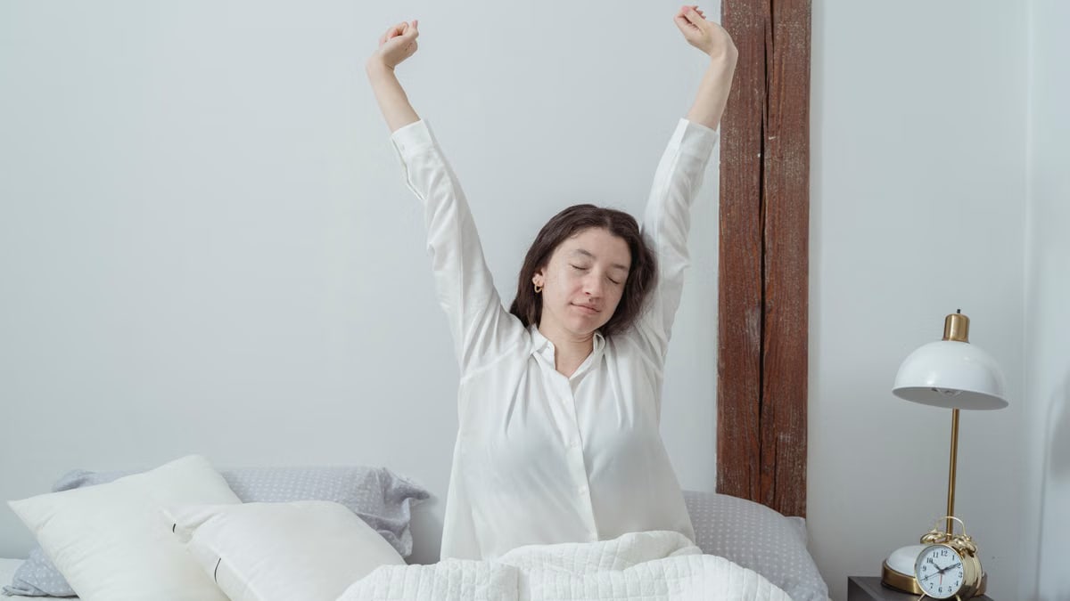 Young brunette with closed eyes sitting on comfortable bed under white blanket and stretching arms up while waking up in morning at home